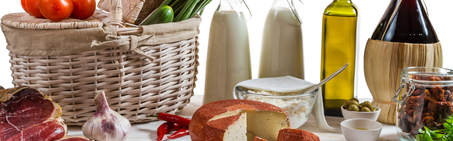 Korb mit regionalen Spezialitäten: Käse, Milchflaschen, Olivenöl, Wein, Schinken, Knoblauch, Oliven und getrocknete Tomaten.